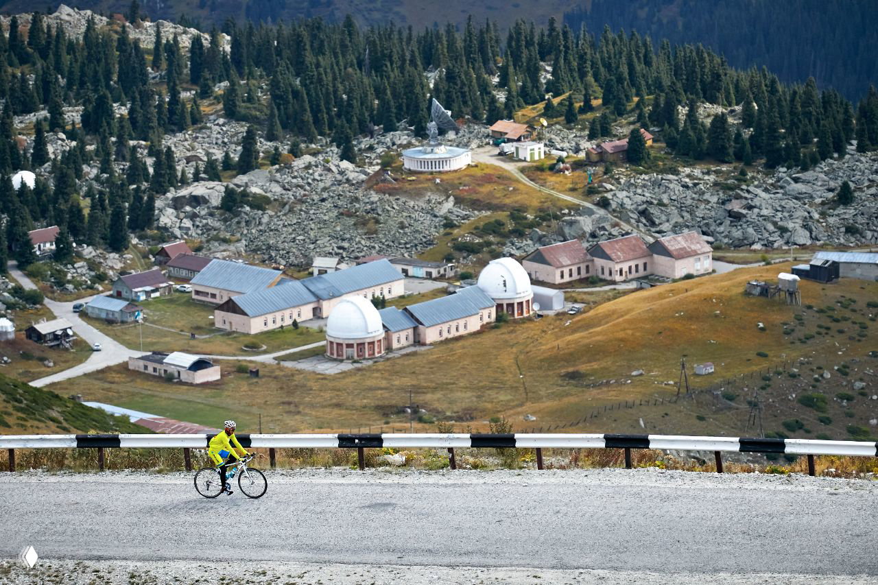 Велосипедист на горной дороге на фоне обсерваторий и хвойного леса; кадр иллюстрирует веломаршрут и подъёмы Kosmos UpHill.