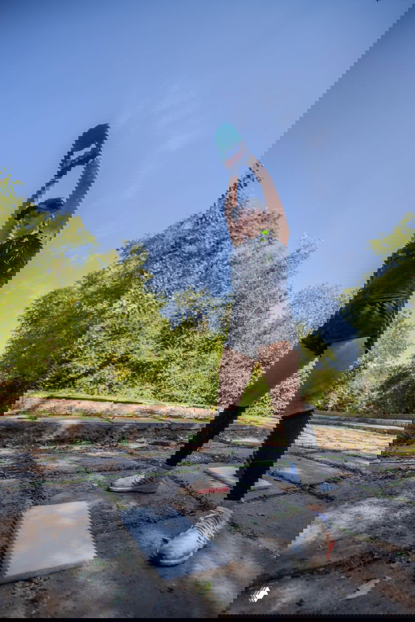 Атлетка поднимает спортивный снаряд над головой на открытой площадке с зелёным фоном, демонстрируя силу в упражнениях городского OCR-старта.