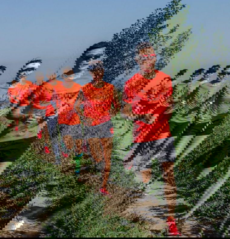 Парк-трейл: группа бегунов в оранжевых футболках бежит по пересечённой грунтовой тропе среди зелёной растительности.