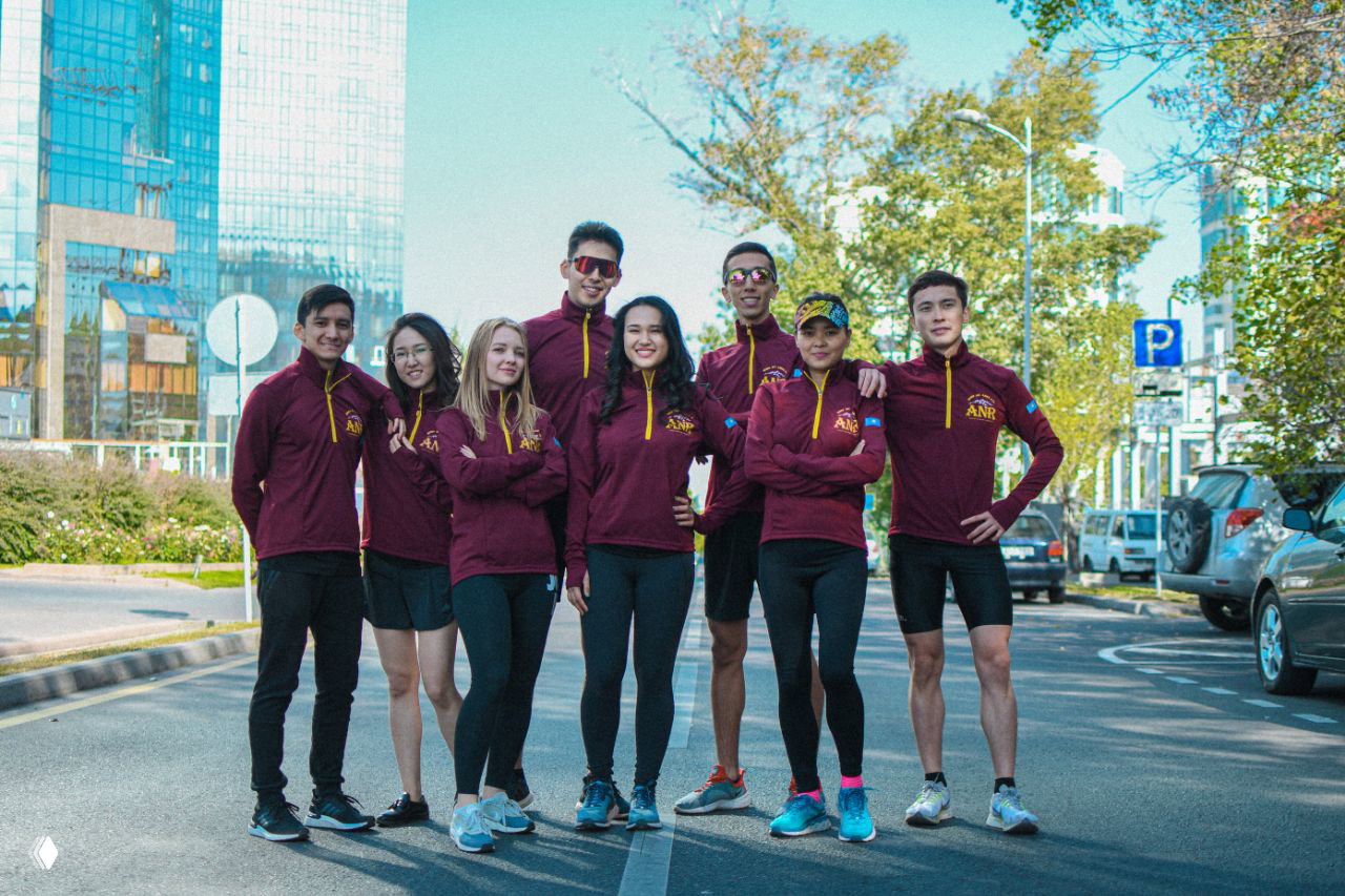 Командное фото бегового клуба ANR на улицах Алма‑Аты: спортсмены в бордовых толстовках, небоскрёбы и автомобили на заднем плане, командный портрет.