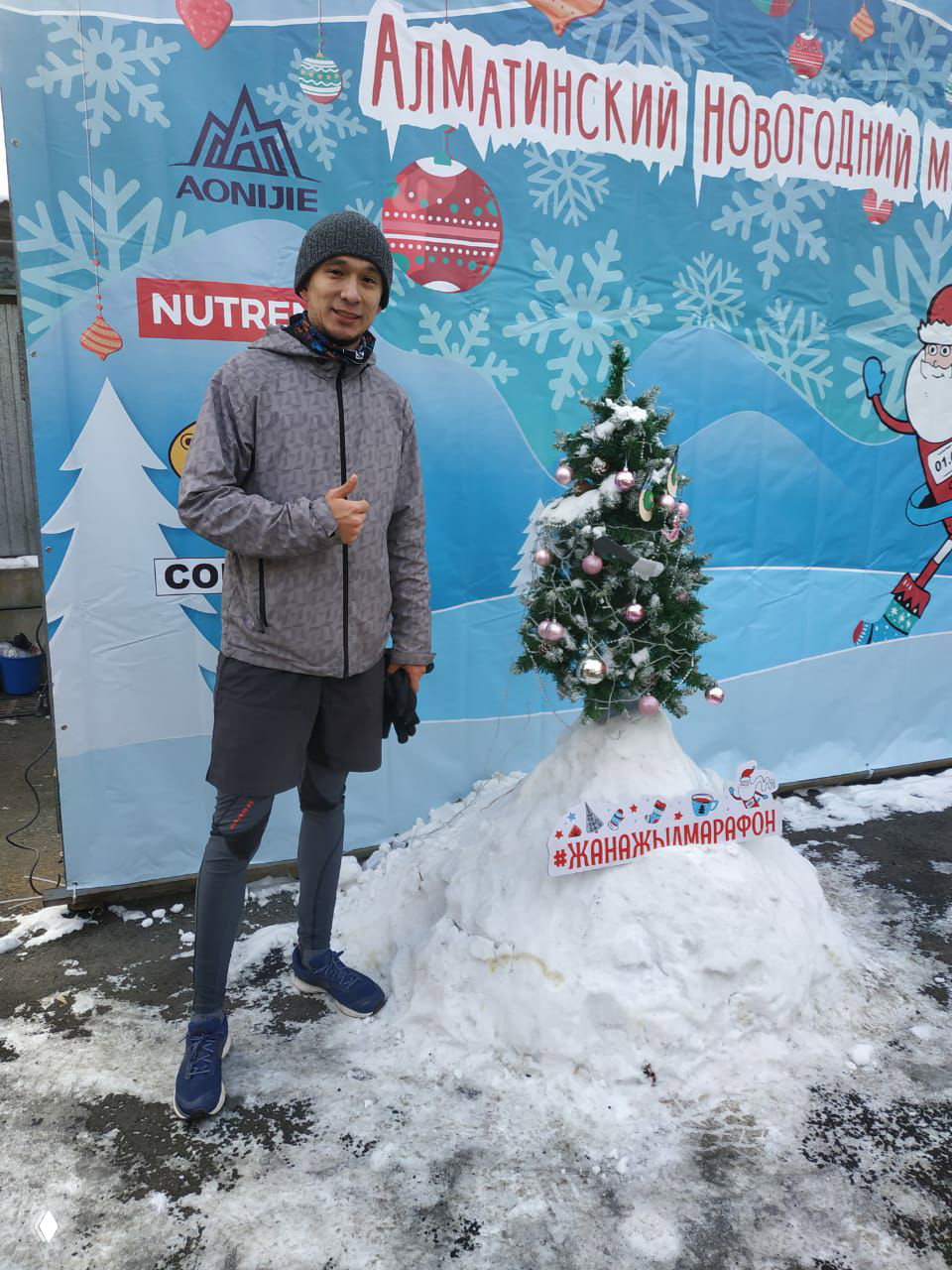 Участник у фото-зоны и маленькой украшенной ёлки на точке выдачи медалей Алматинского Новогоднего марафона, позирует в беговой экипировке на снегу.