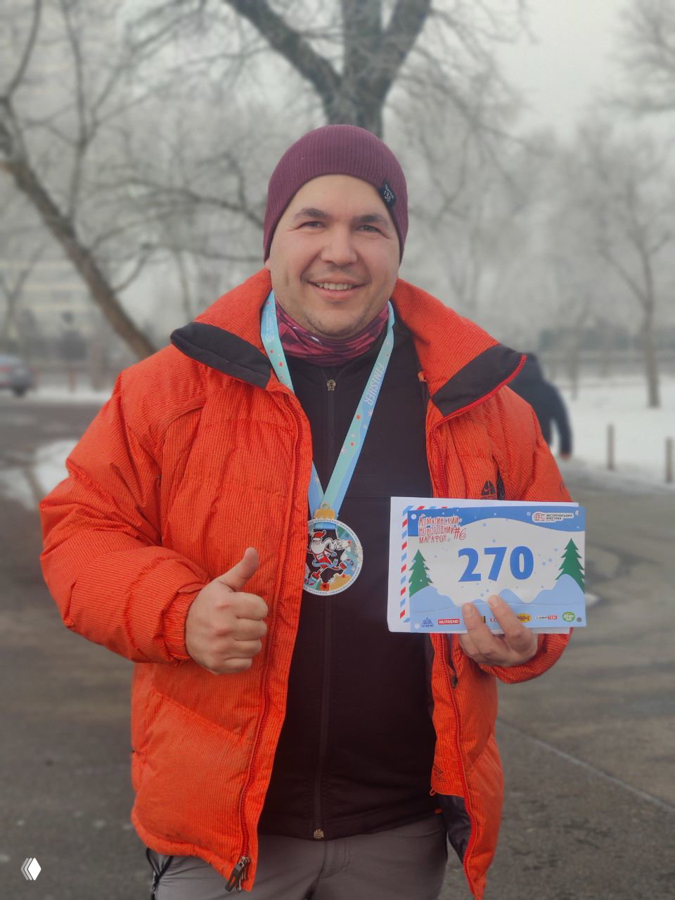 Улыбающийся финишёр в оранжевой куртке показывает медаль на шее и держит табличку с номером 270 после финиша Алматинского Новогоднего марафона, праздничная атмосфера.