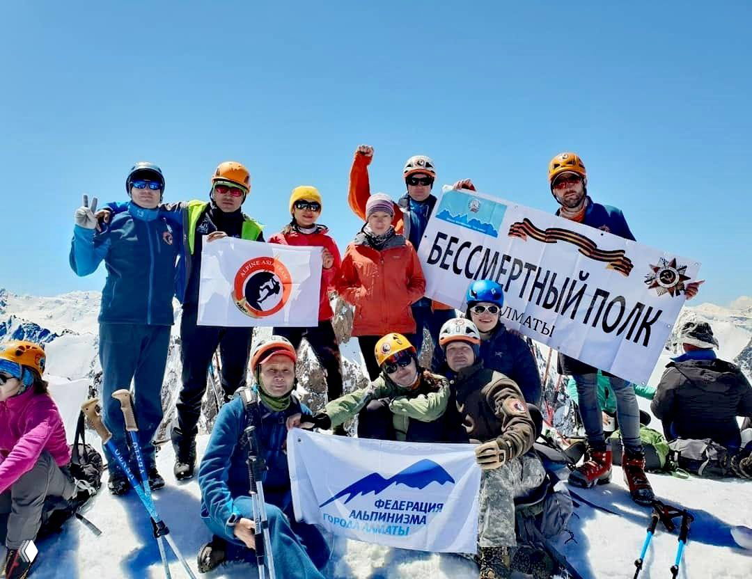 Группа альпинистов на вершине с флагами и баннерами — виден флаг Alpine Asia Team и баннер «Бессмертный полк», снежные пики на фоне ясного неба.