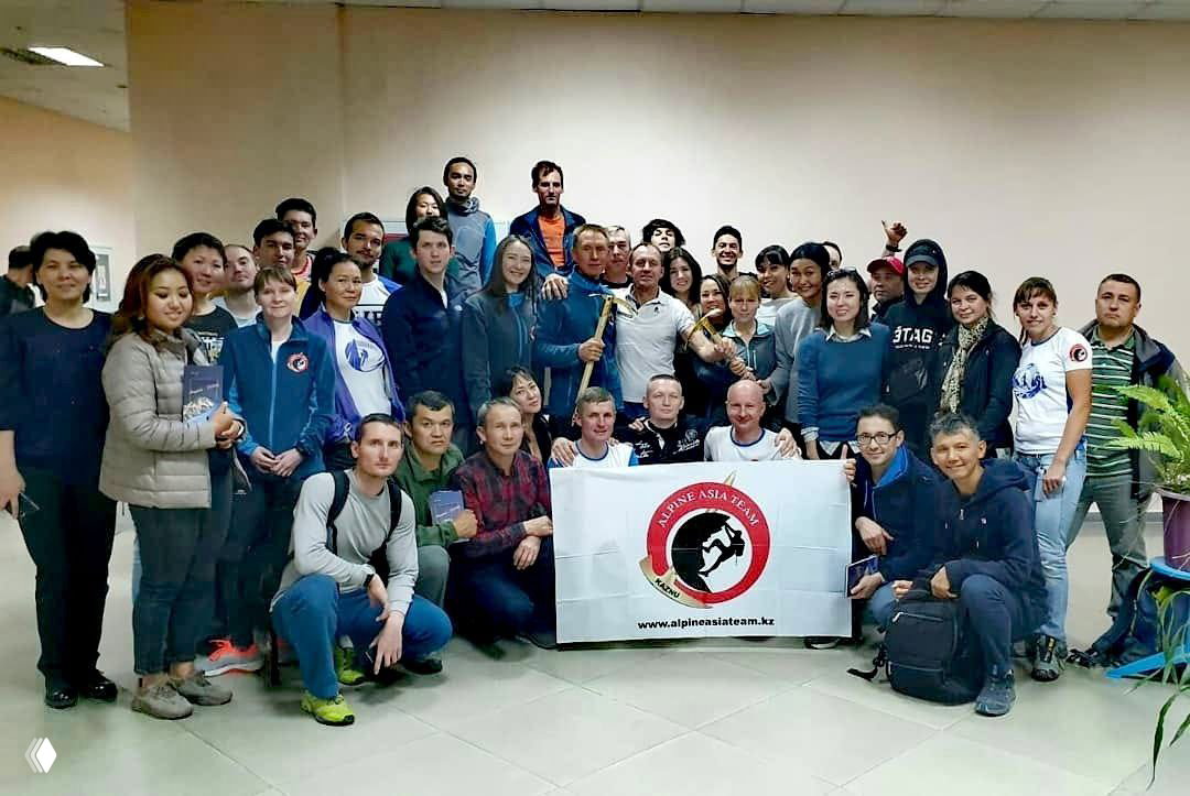 Широкая командная фотография в помещении: участники клуба держат флаг Alpine Asia Team на собрании и командной встрече после мероприятий.