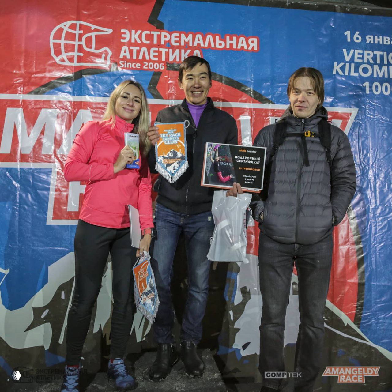 Три человека на подиуме держат вымпелы и призы перед баннером «Экстремальная атлетика» после награждения Amangeldy Race.