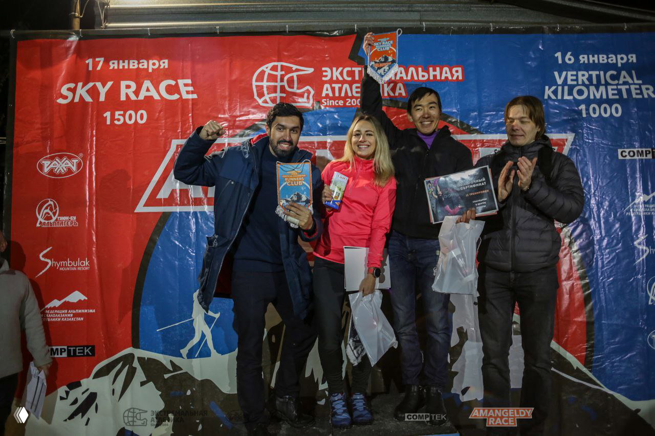Группа участников на сцене с призами и подарочными сертификатами на фоне красно‑синего баннера Amangeldy Race в вечернее время.