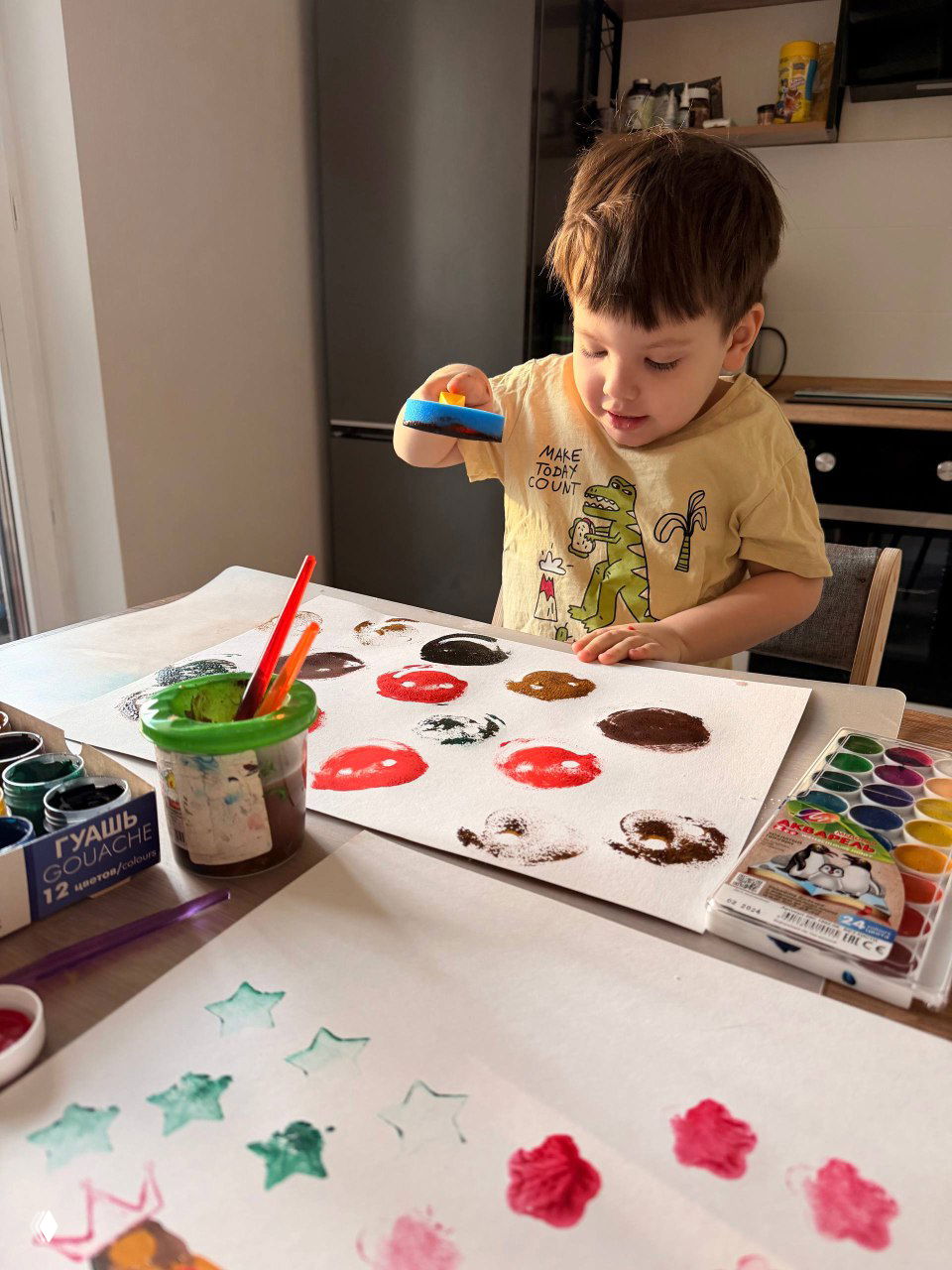 Маленький мальчик сидит за столом и печатает губкой цветные круги на листе бумаги — игровой творческий процесс в домашней кухне.