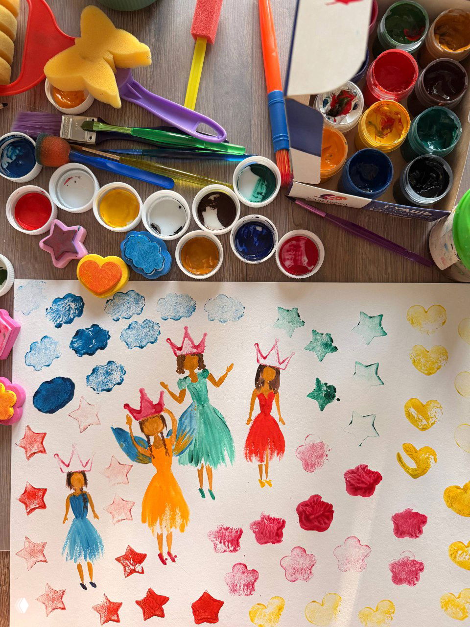 Баночки с краской, кисточки и формы-губки рядом с отпечатками на бумаге — подготовка и результат детской печати в процессе занятия.