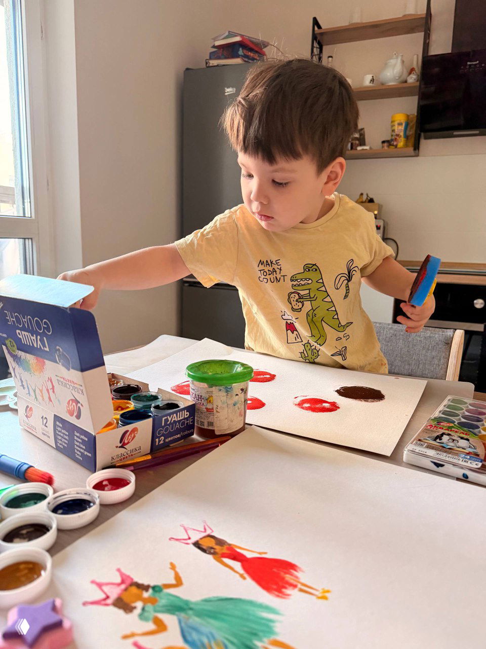 Ребёнок тянется к коробке с гуашью на столе, вокруг листы с отпечатанными звёздами и цветочками — момент домашнего творчества и игры.