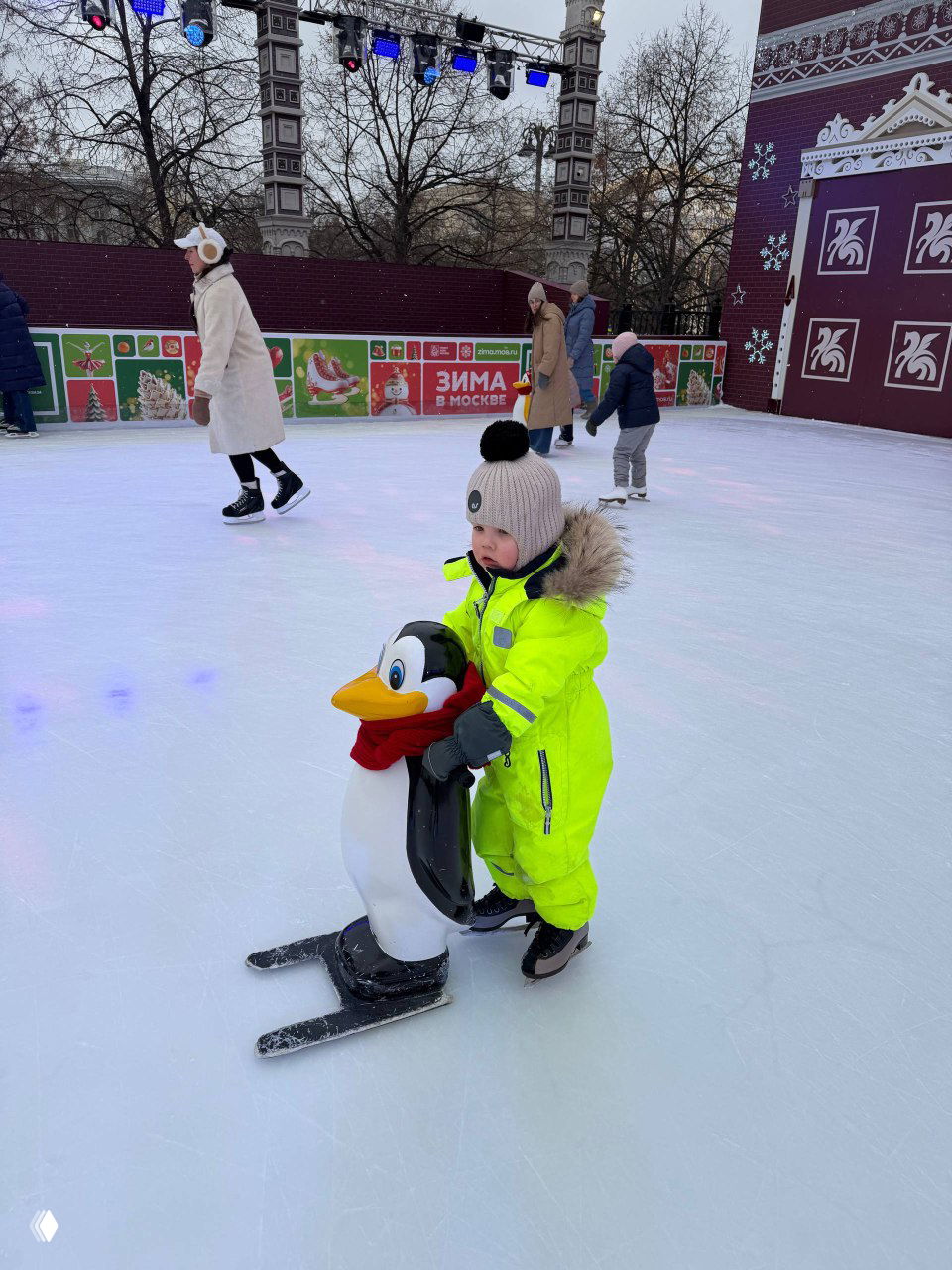 Малыш в жёлтом комбинезоне держится за пластикового «пингвина» на катке, учится кататься на коньках в окружении людей.