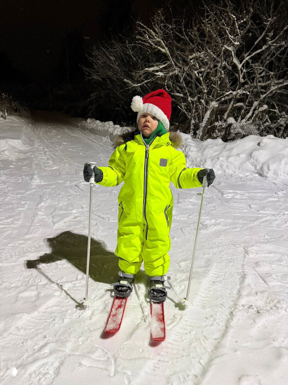 Малыш в неоновом комбинезоне стоит на беговых лыжах ночью, с палками, готовый к прогулке по заснеженному пути.