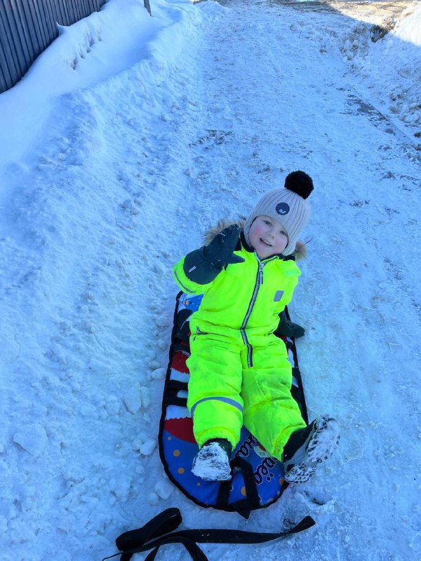 Ребёнок в свете ледяного дня сидит на цветной ватрушке/ледянке и улыбается, готовый съехать по склону во дворе.
