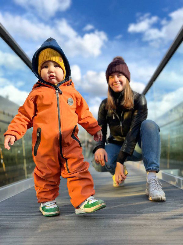Ребёнок в ярком оранжевом комбинезоне идёт по прозрачному пешеходному настилу рядом со взрослым, солнечный день и живые цвета на фото.