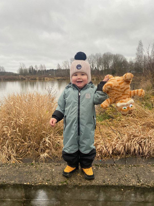 Ребёнок в светло-зелёном комбинезоне стоит на берегу пруда с мягкой игрушкой в руке, осенняя или ранняя весенняя природа вокруг, семейная прогулка.