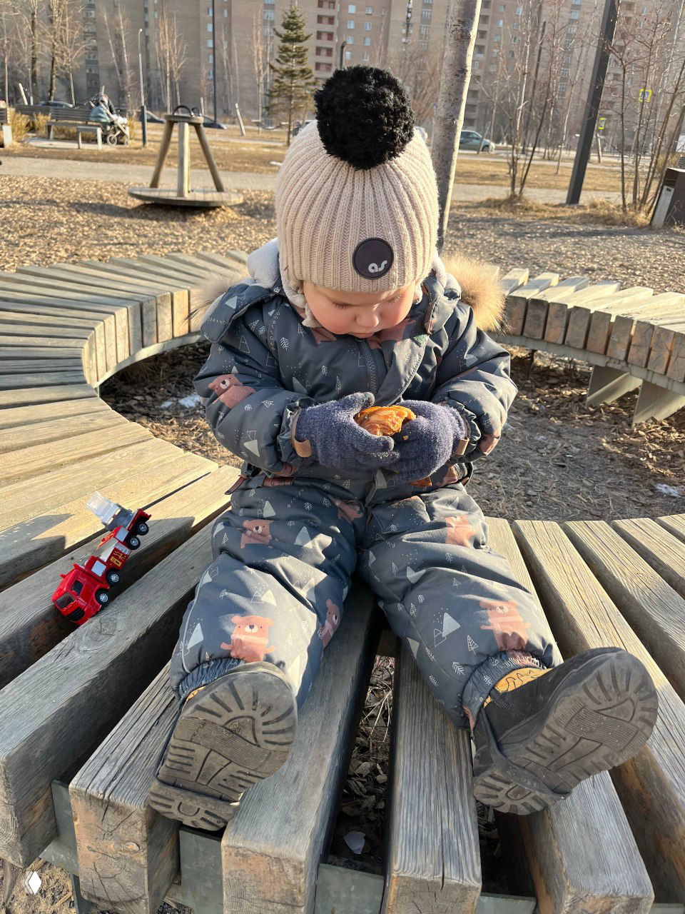 Малыш в зимнем комбинезоне сидит на деревянной скамейке, держит булочку в перчатках, рядом стоит маленькая игрушечная машина — атмосфера дворовой прогулки.