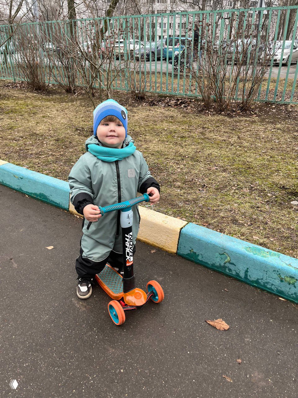 Ребёнок на самокате в светло-зелёном комбинезоне и тёплом шарфе едет по асфальту во дворе, припаркованные машины и кусты на заднем плане, будний день.