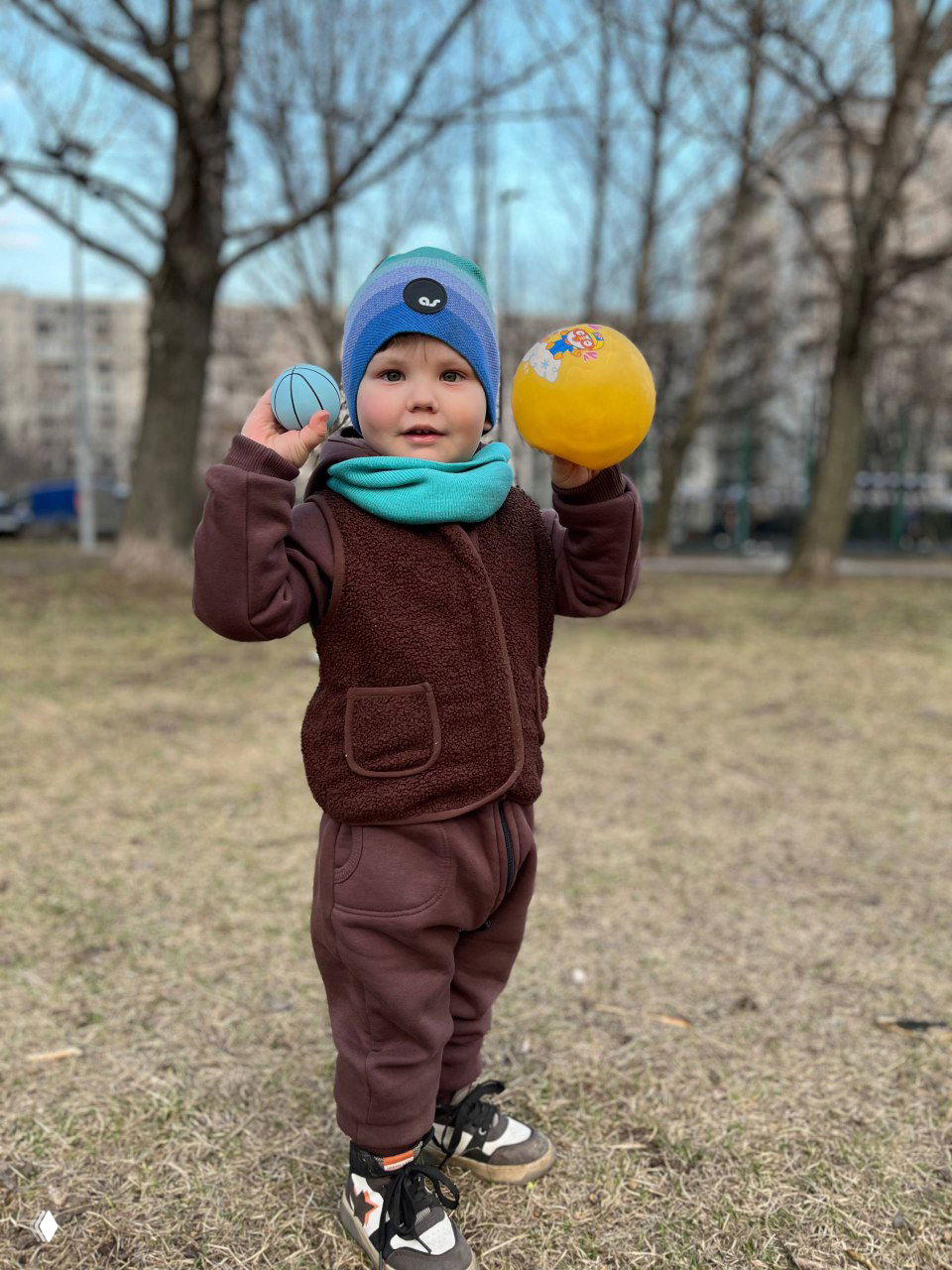 Ребёнок в коричневом комплекте держит два мячика на фоне парка и жилых домов, портрет в полный рост, спокойная игра на свежем воздухе.