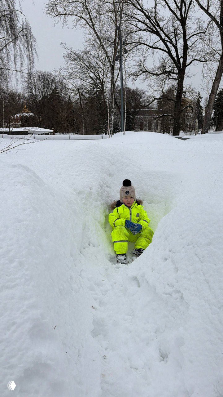 Ребёнок в неоновой жёлто-зелёной зимней одежде сидит в углублении среди высокого снега, глубокий сугроб вокруг и зимний пейзаж, защита от холода.