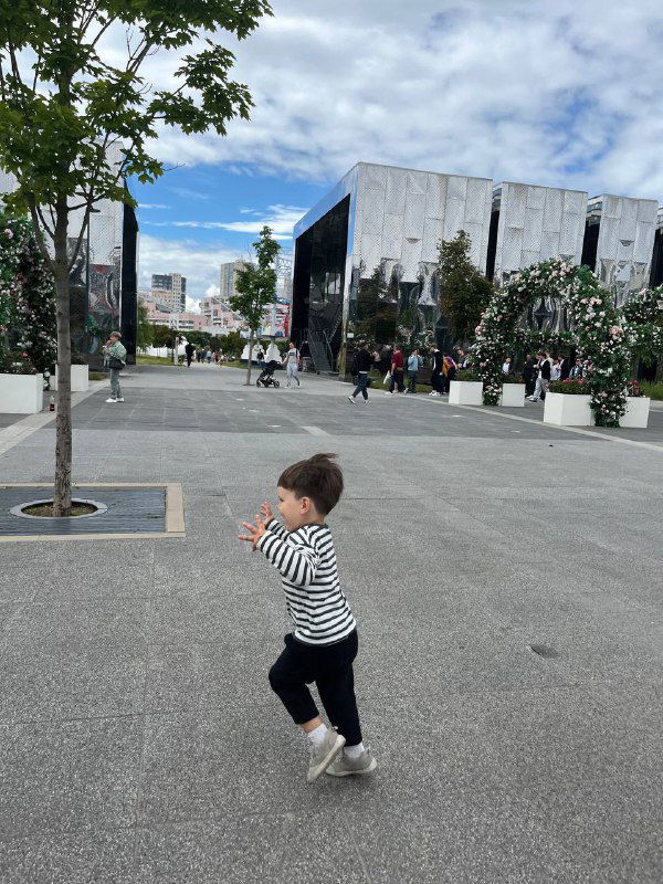 Маленький ребёнок в полосатой кофте бежит по открытой городской площадке; на фоне небо с облаками и архитектура города.