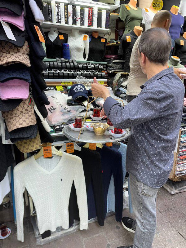 Покупатель в уличной торговой зоне держит в руке стакан чая возле прилавка с одеждой — уличная торговля и чаепитие рядом с товарами.