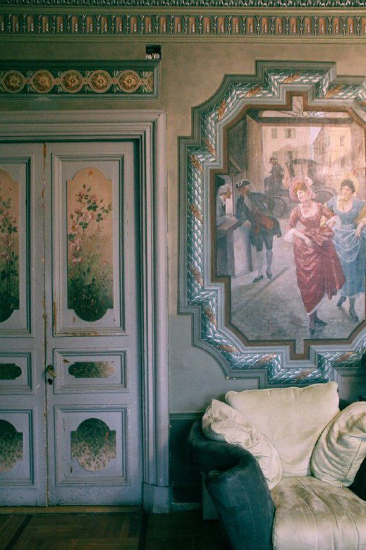 Декоративные панно и двери с росписью в историческом интерьере особняка, уютный диван у стены и изящные молдинги.