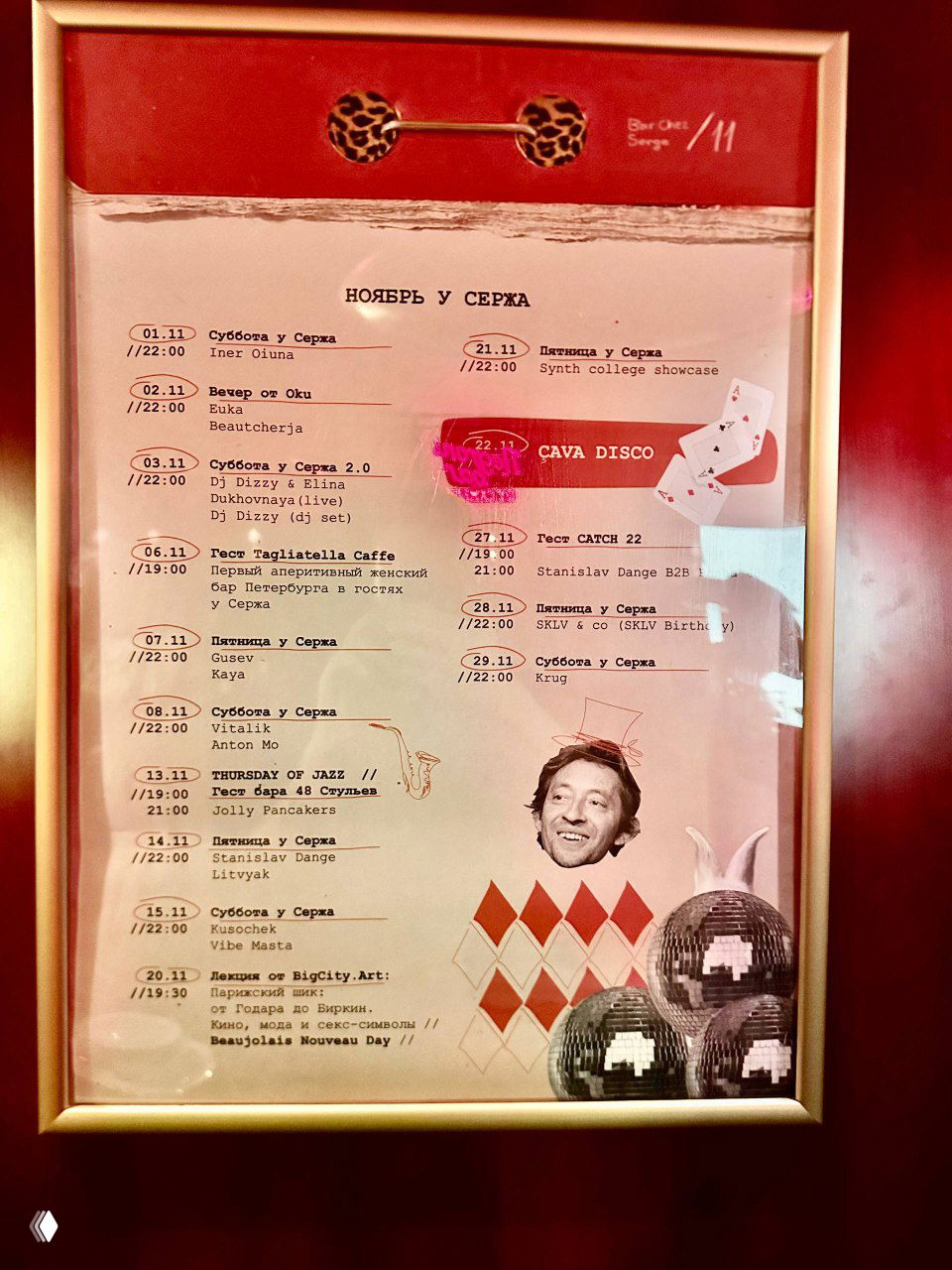 Рамка с афишей мероприятий «Ноябрь у Сержa» в золотой рамке на красной стене — расписание вечеринок и концертов в баре Chez Serge.
