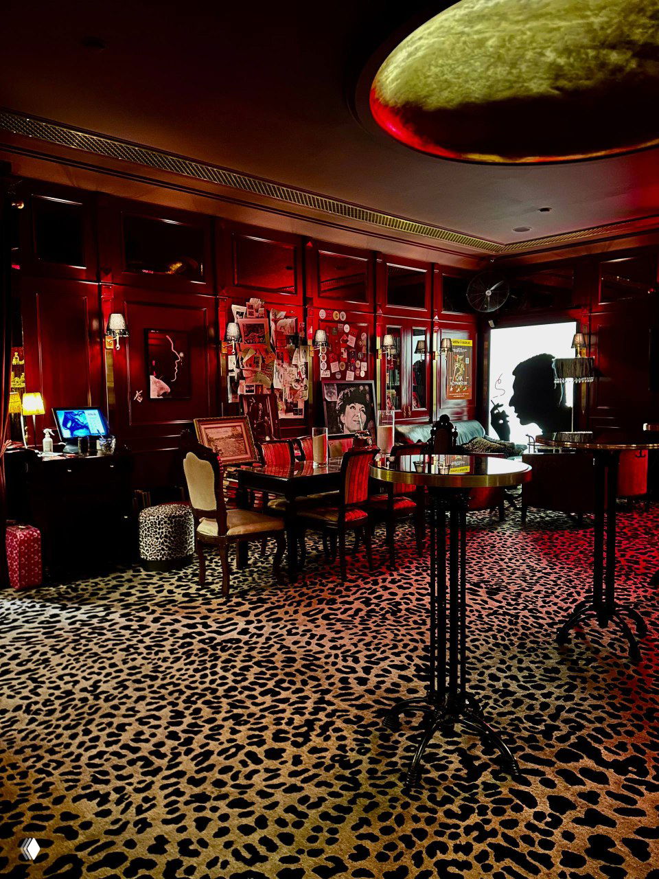 Общий вид зала с низкими столами, лампами и леопардовым ковровым покрытием — тёплый, театральный салонный интерьер Chez Serge в красных тонах.