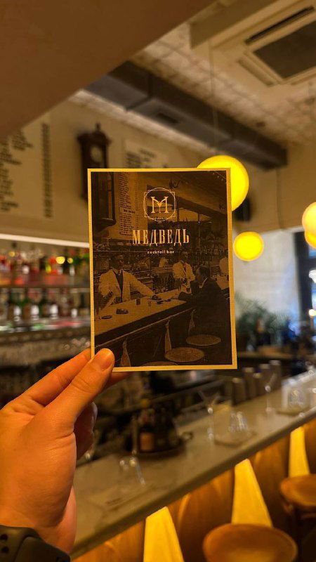 Рука держит карточку меню бара на фоне барной стойки с подсветкой — интерьер современного бара, где проходят встречи и экскурсии.