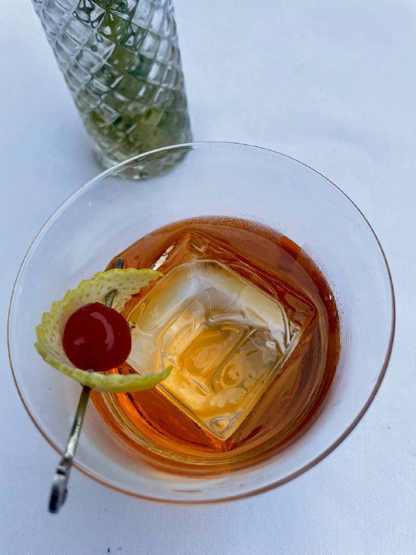 Крупный план коктейля с большим квадратным льдом, вишней и цедрой — вид сверху; интерьерная деталь бара «Медведь».