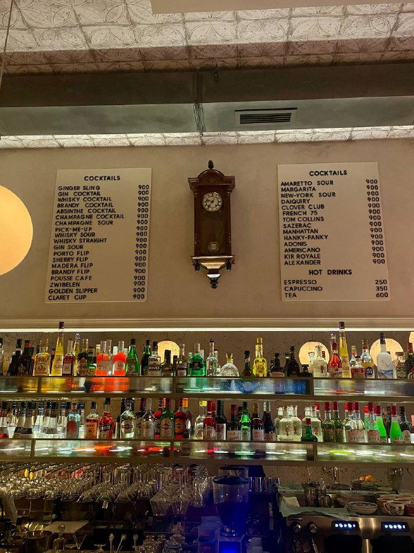 Барная стойка с полками бутылок и старинными часами на стене; на фоне видна большая доска-меню с перечнем коктейлей и цен в «Медведе».