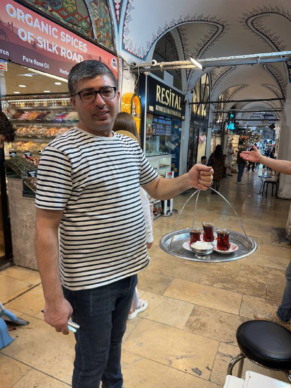 Продавец в полосатой футболке нес поднос с несколькими стаканами чая через крытую торговую галерею — типичная сцена подачи чая посетителям базара.