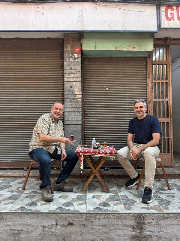 Двое мужчин сидят за маленьким столиком у закрытого магазина на тротуаре, перед ними стаканы крепкого турецкого чая и простая уличная обстановка.