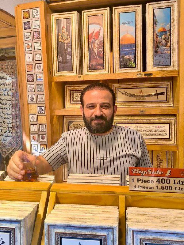 Продавец восточных сувениров или картин позирует за прилавком с чашкой чая — интерьер лавки с рамками и предметами искусства на заднем плане.