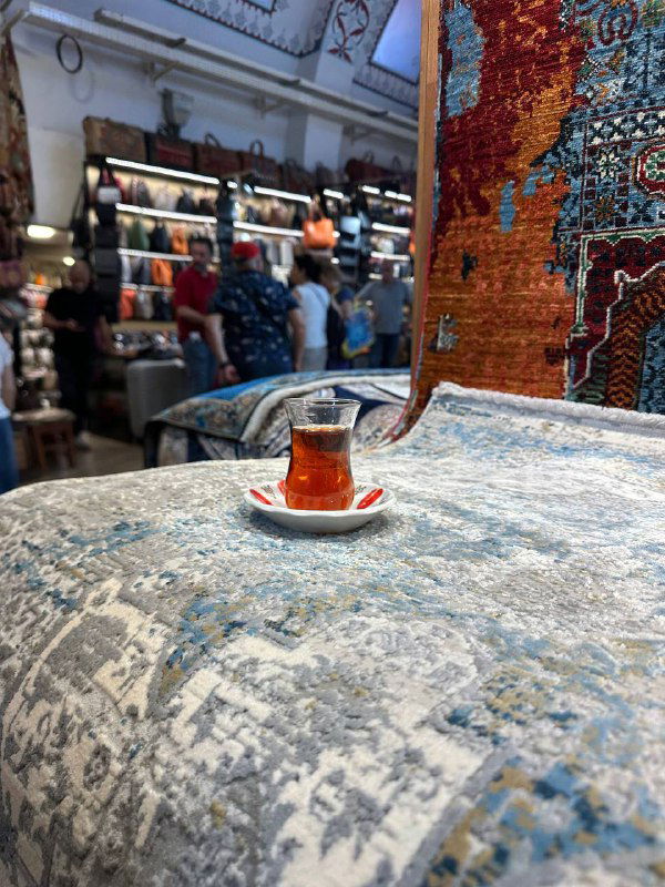 Одинокий стакан чая на столике, поставленном на покрытый ковром стол в магазине ковров; акцент на чашке и текстуре ковра вокруг.