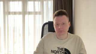 Фотография мужчины в светлой комнате за столом, в майке с логотипом The North Face; нейтральное выражение, окно и шторы на фоне.