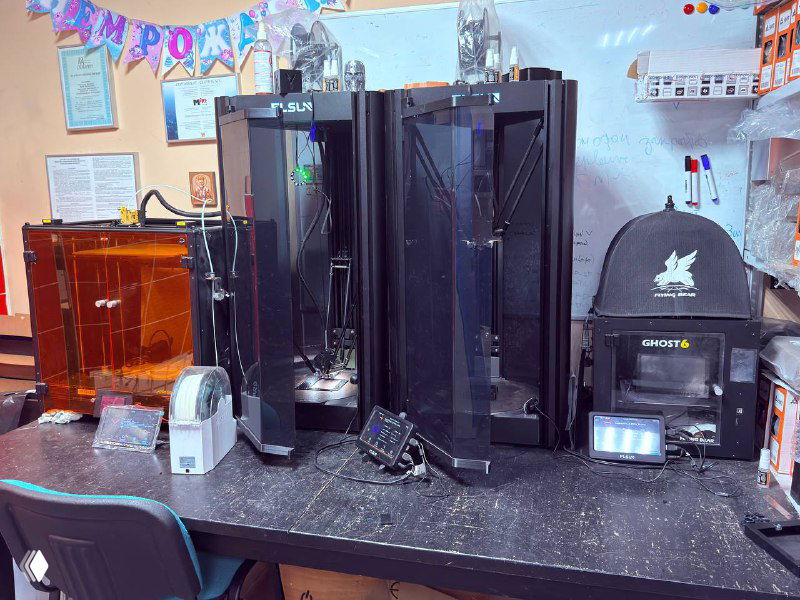 Крупные вертикальные 3D‑принтеры и стол с инструментами в лаборатории — рабочее место инженеров и операторов печати.