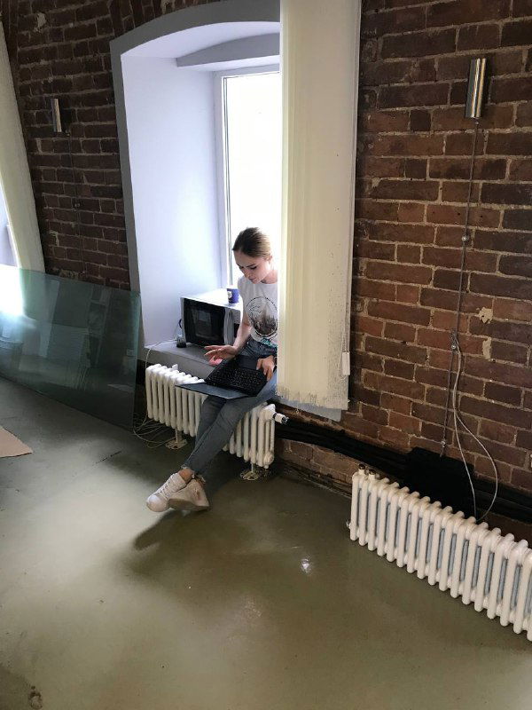 Человек с ноутбуком сидит на подоконнике у батареи в ещё неготовом офисе с кирпичной кладкой и следами ремонта — временное рабочее место.