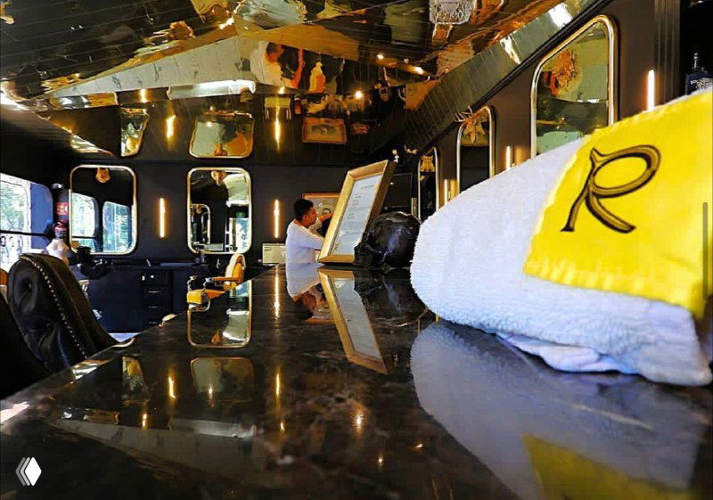 Интерьер барбершопа с полированными тёмными поверхностями, полотенцем с буквой R и рабочими местами на переднем плане; акцент на деталях оформления.