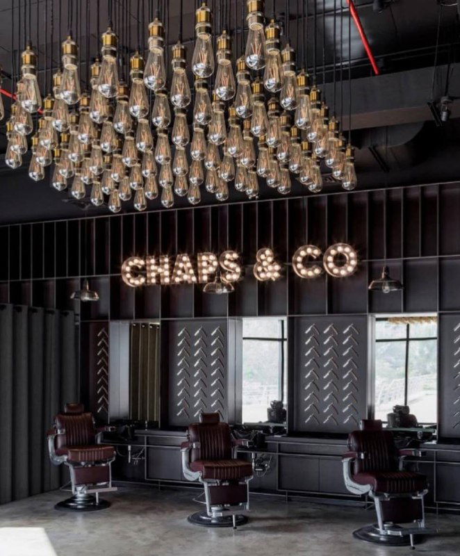 Зал Chaps &amp; Co с винтажными креслами и крупной люстрой из подвешенных ламп; индустриальный тёмный интерьер и светящаяся вывеска бренда над помещением.