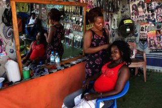 Внутренний вид салона в Кении: мастерица заплетает волосы клиентке, вокруг — плакаты с примерами причёсок и уходовые средства.