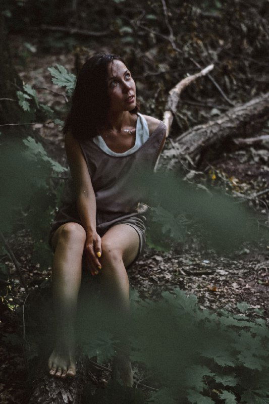 Девушка сидит на земле в лесу, взгляд в сторону, мягкий свет и размытый передний план создают ощущение интимного размышления и внутренней честности.