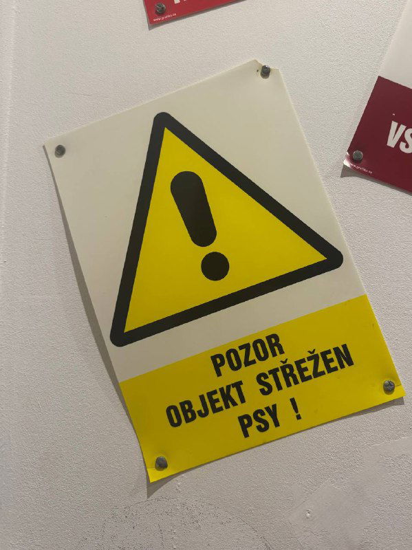 Жёлтая сигнальная табличка с треугольником и предупреждением на иностранном языке, прибитая к стене — элемент интерьера или декора.