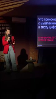Лектор держит микрофон на тёмной сцене; свет фокусируется на спикере, слайд с крупным текстом лекции виден позади, аудитория в тени.