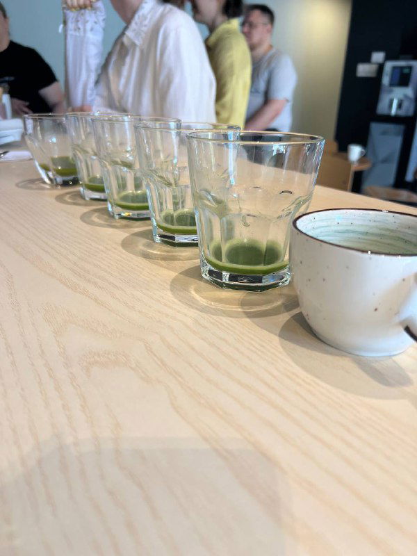 Ряд прозрачных стаканов с остатками зелёного порошка матчи на дне, на переднем плане белая чашка, фон с людьми на дегустации слегка размыт.