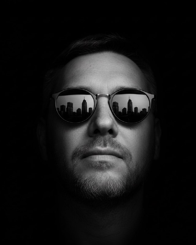 Контрастный чёрно‑белый портрет мужчины в солнцезащитных очках с отражением городской панорамы в линзах, стилизованное изображение.