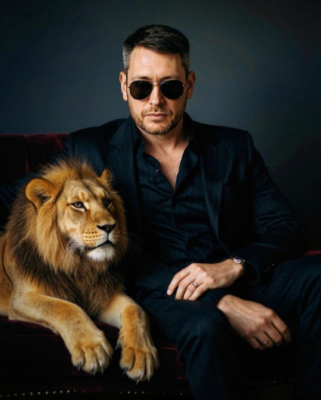Мужчина в тёмном костюме рядом с львом — стилизованный кинематографичный портрет, подчёркивающий силу и композицию кадра.