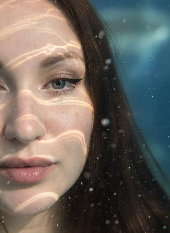 Макро‑портрет женщины в воде: левая часть лица под поверхностью, мягкие тени и яркие блики каустики, создающие мечтательную атмосферу.
