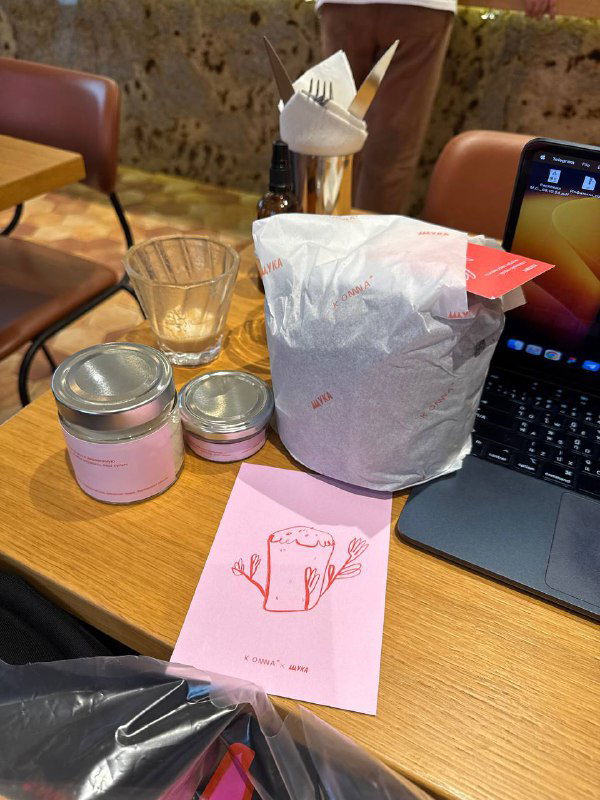 Пасхальный набор кофейни на столе в кафе рядом с ноутбуком: баночки, бумажный пакет и розовая карточка с иллюстрацией, рабочая атмосфера.