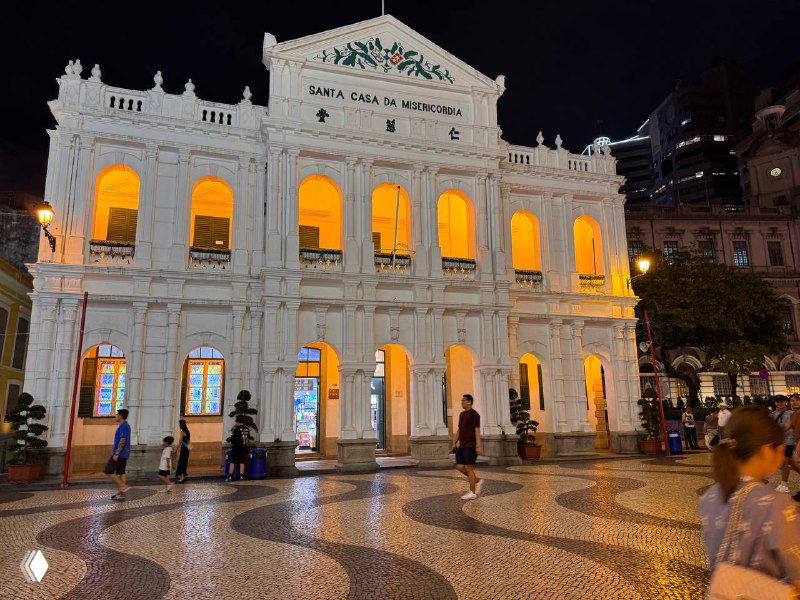 Белое колониальное здание подсвечено ночью, перед ним характерная мозаичная португальская брусчатка и прохожие — типичная сцена старого Макао.