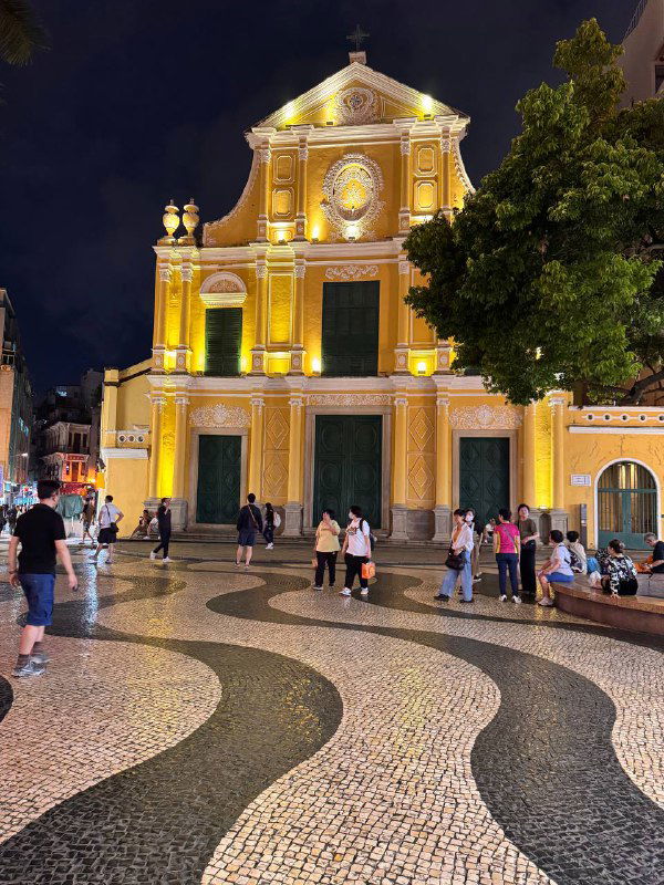 Фасад освещённой церкви у площади с волнообразной плиткой и гуляющими людьми — сочетание религиозного памятника и туристической площади в Макао.